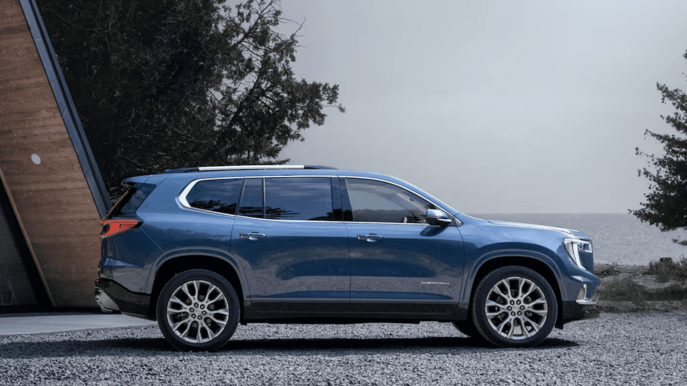 Side profile of the 2025 GMC Acadia Denali in blue, parked near a modern cabin and lake in West Jefferson, NC.