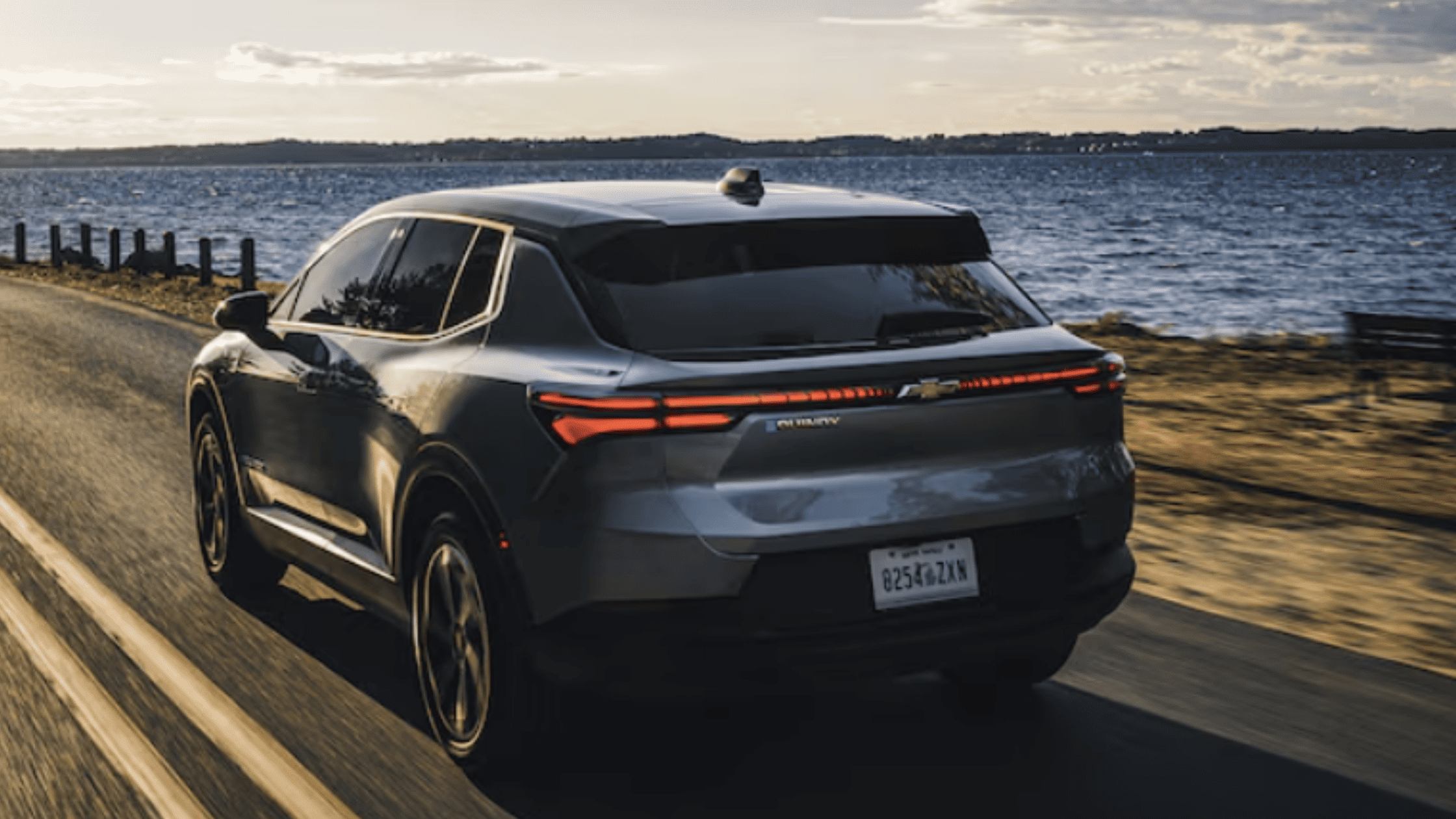 Rear three-quarter view of the 2026 Chevrolet Equinox EV driving along a waterfront road, showing sleek taillights and aerodynamic electric SUV design.