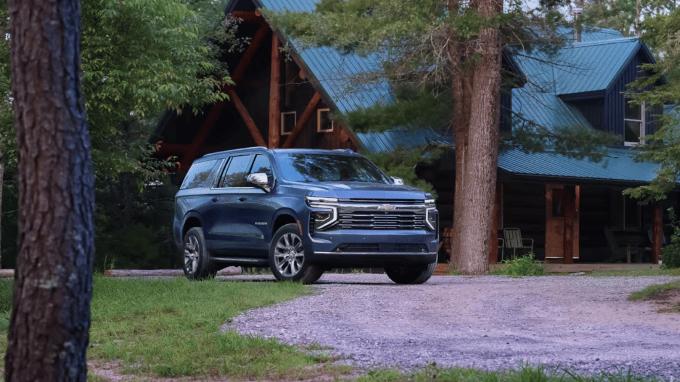 2026 Chevrolet Suburban vs Ford Expedition for Morganton NC Drivers With Growing Families – blue Chevrolet Suburban SUV parked in front of a wooded cabin, showcasing full-size SUV space and family travel appeal.
