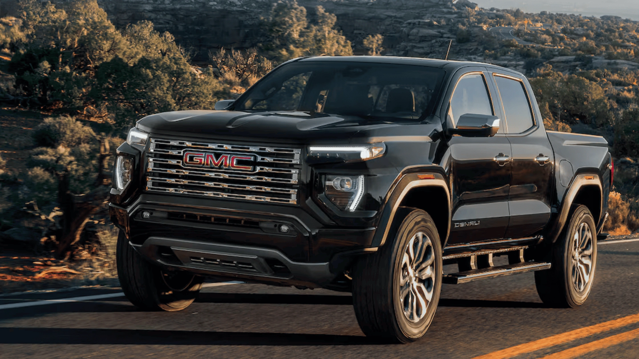 2026 GMC Canyon Denali midsize pickup truck in black with chrome grille and Denali trim driving on desert highway in Boone NC