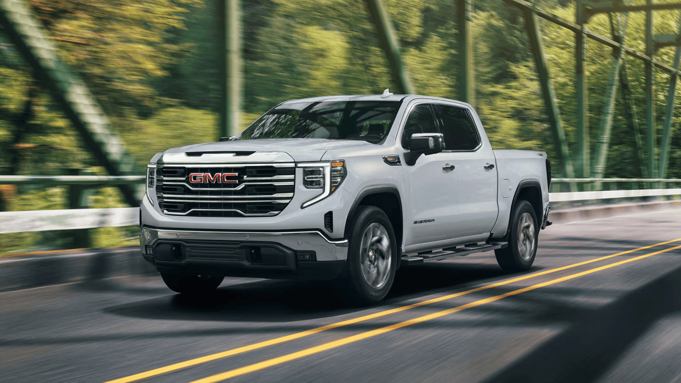 2026 GMC Sierra 1500 crew cab pickup truck in white driving across bridge surrounded by lush green trees in Boone NC