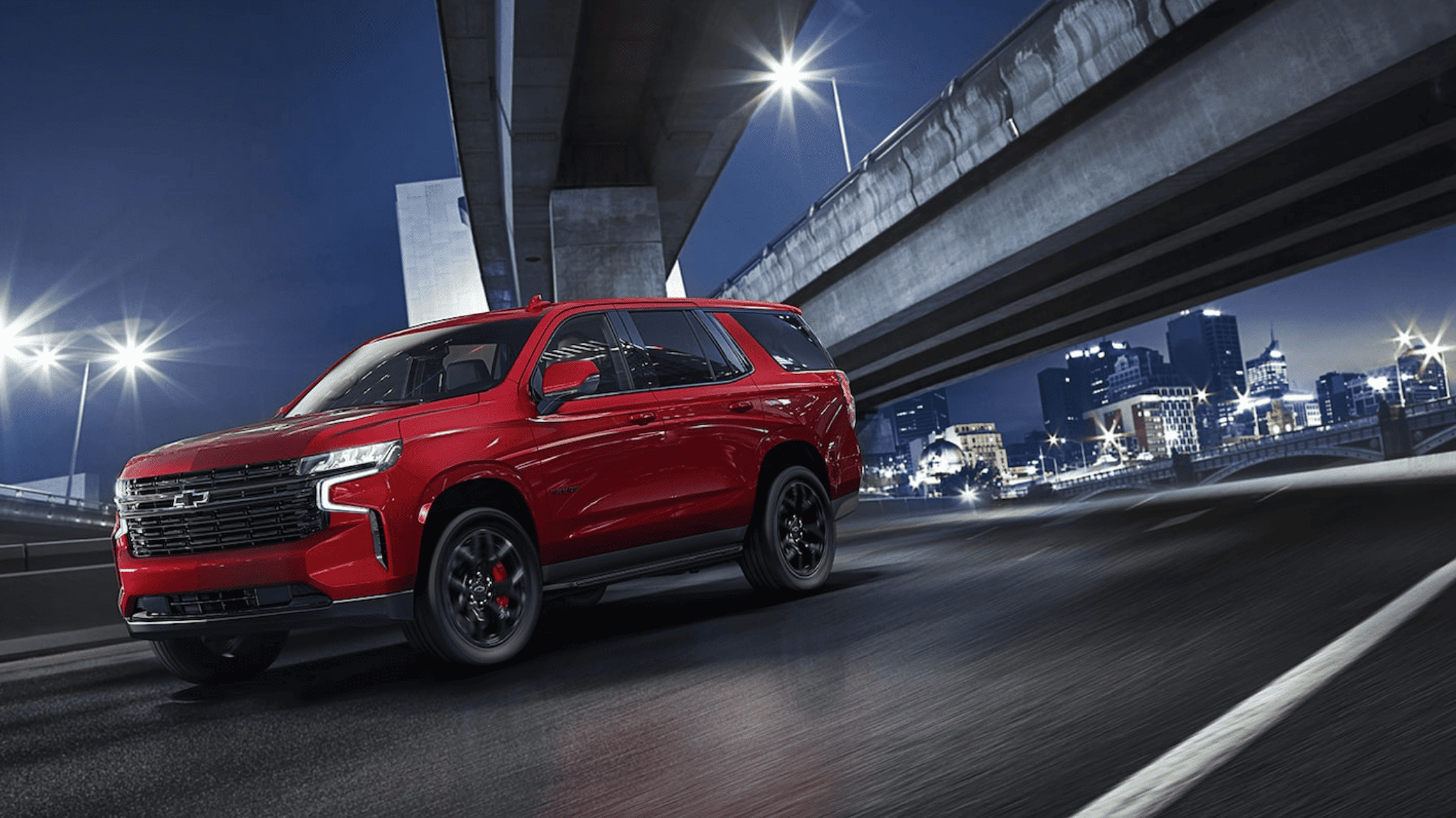 Front side view of red Chevrolet Tahoe RST driving under city overpass.