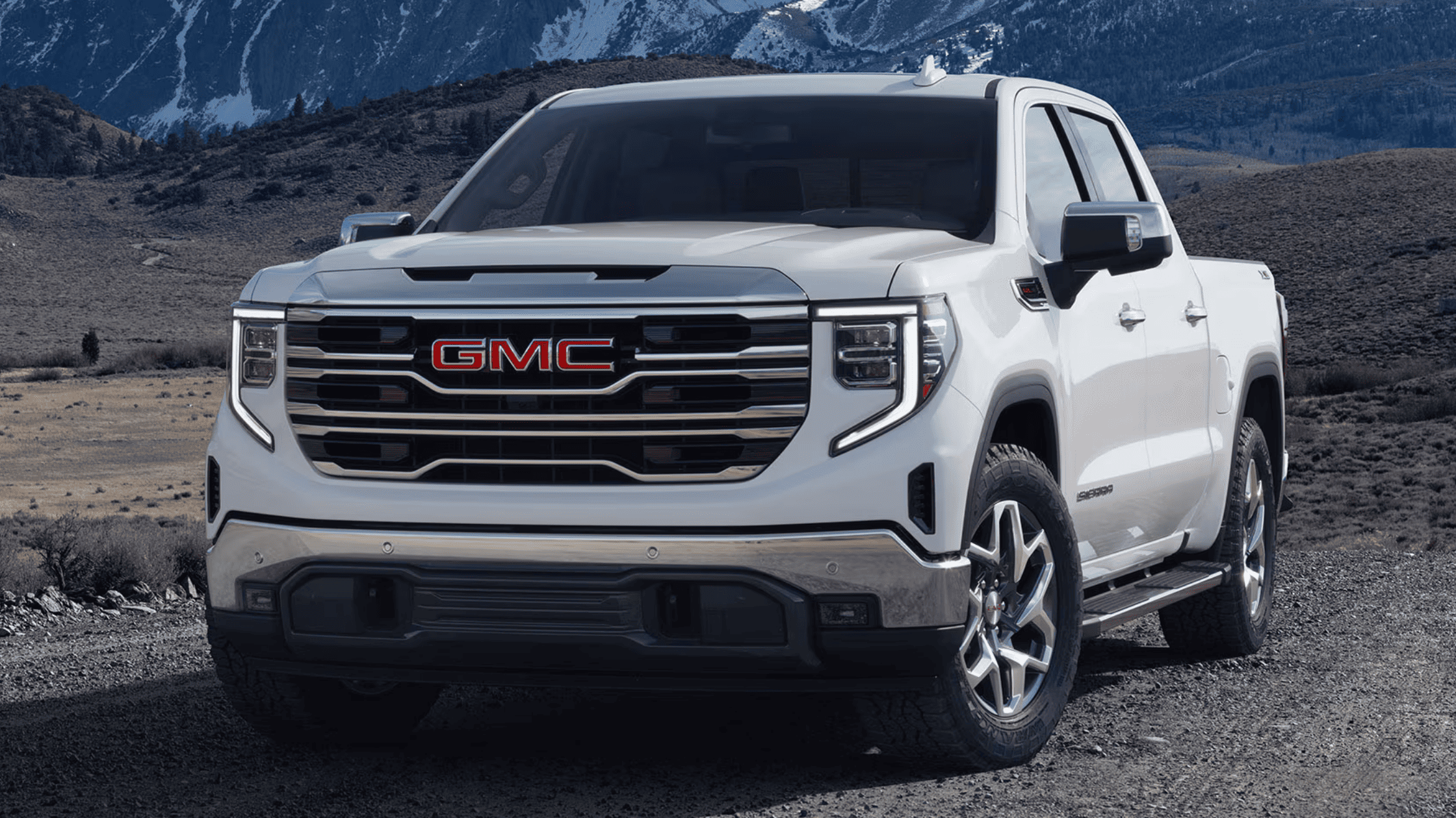 Front angle of the 2025 GMC Sierra 1500 in white, showcasing its bold grille and chrome accents in a mountainous terrain.