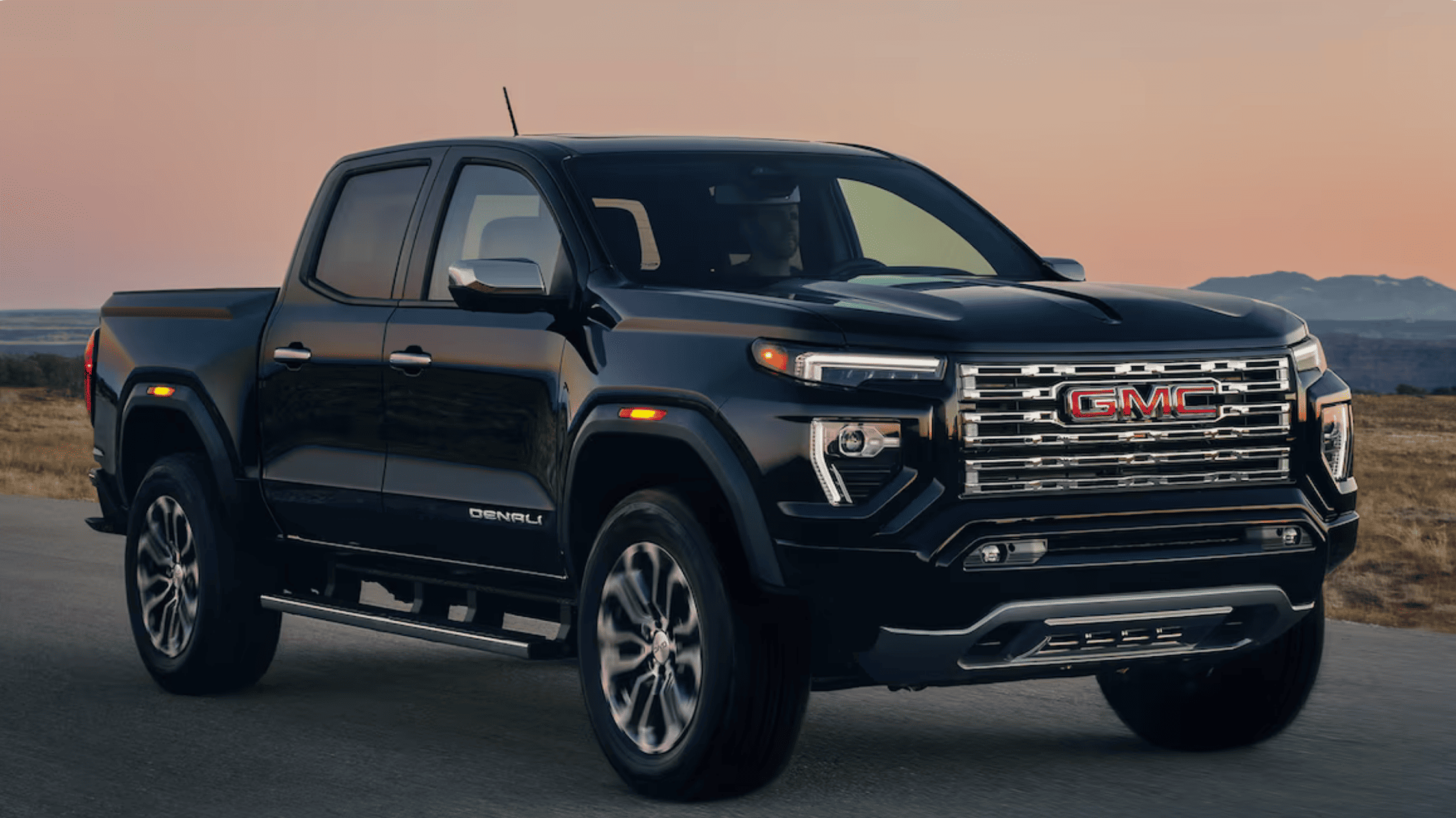 Black 2026 GMC Canyon pickup truck driving on a scenic road in Boone, NC during sunset, showcasing rugged performance and design.